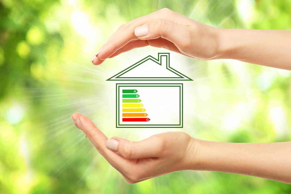 Mâinile unei femei înconjurând desenul unei case cu imaginea unei scale de eficiență energetică pe un fundal natural